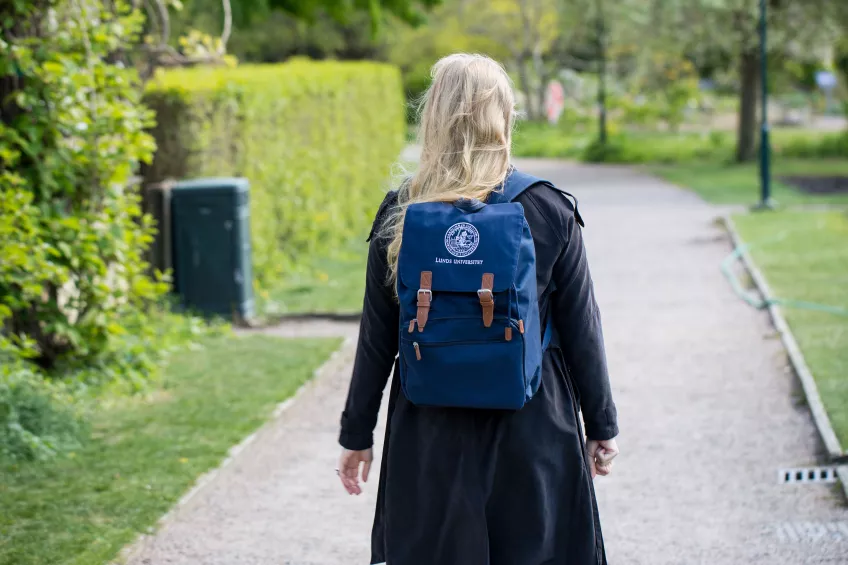Studentska med Lunds universitet-ryggsäck promenerar i park. Foto.