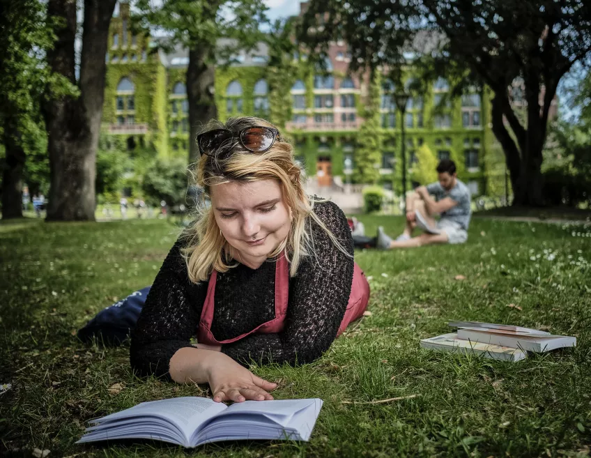 Foto av en kvinnlig student som ligger och läser i gräset utanför Universitetsbiblioteket.